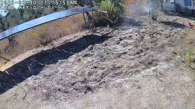 A fixed camera at the camp: a long ground-mounted solar array on the left, freshly dug earth, a hose, a white caravan at the top right, under hard midday sun